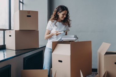 Curly young woman, student is housing in residential room. Happy european woman unpacking boxes with books. Girl in white t-shirt indoors. Relocation and independence concept.
