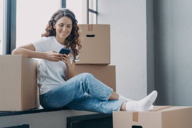 Happy spanish woman is sitting on windowsill of new home. Girl is unpacking boxes. Young lady is texting on phone and smiling. Joy of relocation concept.