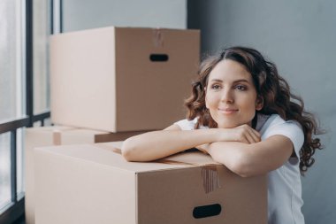 Happy attractive european woman unpacking things and being leaned to boxes. Girl is dreaming near window while sitting on floor in new apartment. Concept of future and perspective.