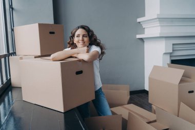 Young lady is dreaming while sitting on floor in new home. Happy attractive hispanic woman unpacking boxes. Large living room with big window and fireplace. Concept of future and perspective.