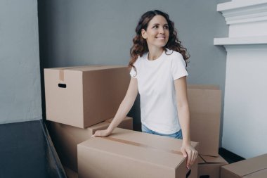 Hopeful lady is holding cardboard box and moving to modern apartment. Happy european woman purchasing real estate and looking at interior. Improvement, changes and new life concept.