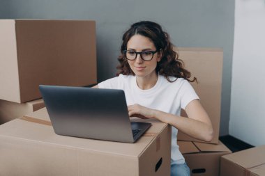 Girl in white t-shirt and glasses is texting on computer ordering delivery service. Happy hispanic woman packing boxes for moving to new place. Easy move concept.