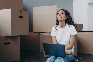 Hopeful hispanic woman sitting among cardboard boxes and holding pc. Lady is in waiting for shipping service and chatting online. Romantic relationship and relocation concept.