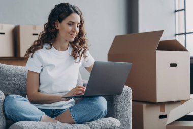 Hispanic girl sitting with laptop among cardboard boxes and smiling. Happy young woman using internet in new home. Lady moves to modern apartment. Relocation and online work concept.