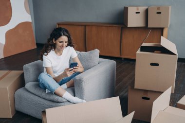 Young spanish woman posting news. Happy apartment buyer unpacking boxes. Girl is moving to new home. Relaxed young lady clicks mobile phone. Independence and achievement concept.