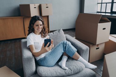 Young european woman posting news and smiling. Happy apartment buyer among boxes. Satisfied young woman is moving to new home. Girl clicks phone and chatting. Independence and achievement concept.