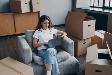Happy young hispanic woman renting apartment online. Girl among cardboard boxes is using laptop and smiling. Happy homeowner is sitting in armchair with pc. Woman about to move.