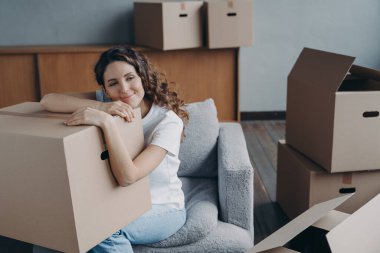 Hispanic girl relocates to new home. Lady is sitting in armchair among packed cardboard boxes. Woman is dreaming leaned to box. Happy young woman moves alone. Independence and success concept.