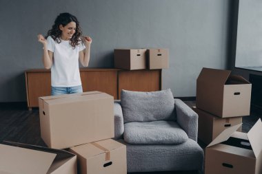 Excited young woman is packing things. Spanish girl moves to new house. Lady is unpacking cardboard boxes in new apartment. Big new living room with furniture. Real estate purchase concept.