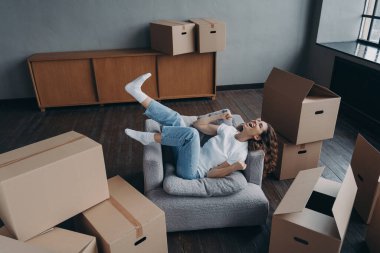 Overjoyed young woman has bought a new apartment. Happy hispanic lady relaxing in armchair. Pretty spanish girl at home with boxes. Concept of opportunity for woman. Independence and new lifestyle.