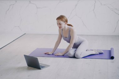 Young caucasian woman practicing yoga on mat on floor of her room. Internet fitness lesson with computer on self isolation. Concept of e-learning and home online classes on corona virus quarantine.