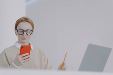 Happy young woman is chatting on phone while studying. Girl is sending messages while holding pencil. Browsing through internet, having fun and smiling. Being distracted at online study concept.