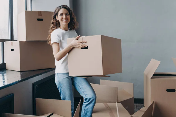 Girl in jeans and white t-shirt is carrying cardboard box. Happy attractive european woman packing things to move. Packed boxes on the floor. Relocation and moving to new apartment concept.