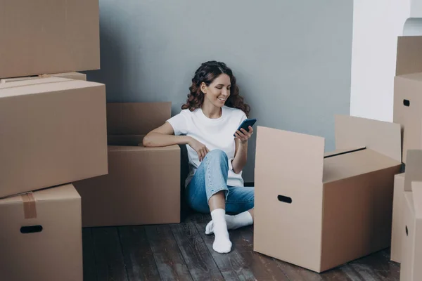 Happy spanish woman getting message from shipping service. Lady is packing boxes for the relocation. Girl is chatting on mobile phone and sitting on the floor. Easy moving concept.