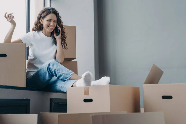 Happy hispanic woman is sitting on windowsill of new house. Girl is unpacking boxes and talking on phone to friends. Relocation and communication concept.