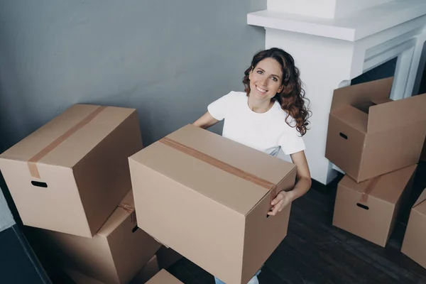 Attractive european girl in white t-shirt is leaving house. Lady is packing cardboard boxes and carrying. Cheerful young woman is going to buy apartment. Relocation and independence concept.