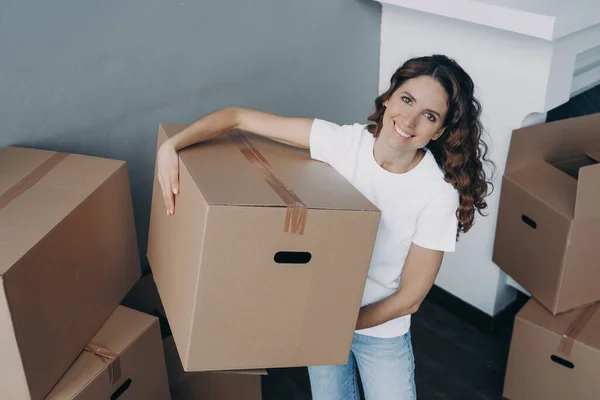 Happy hispanic woman purchasing real estate and carrying box. Lady is unloading cardboard boxes alone in new apartment. Young lady is going to rent house. Mortgage loan and investment concept.