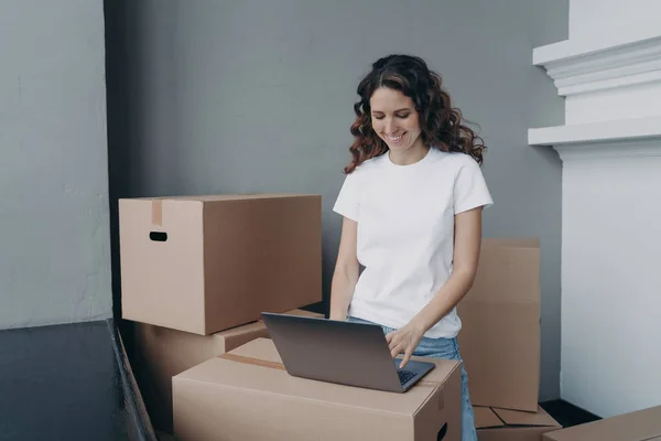 Happy european woman unloading cardboard boxes at new place. Girl in white t-shirt is typing on laptop on the box. Relocation and distance work concept.