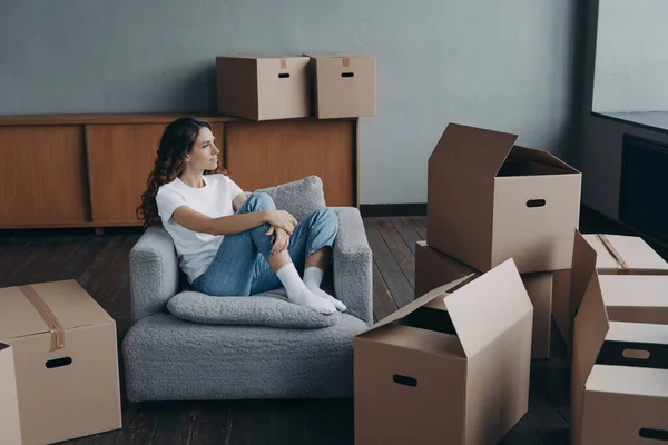 Happy woman is an apartment buyer. Satisfied spanish girl is sitting in armchair. Mover is dreaming among boxes in living room of new home. New house owner having rest.