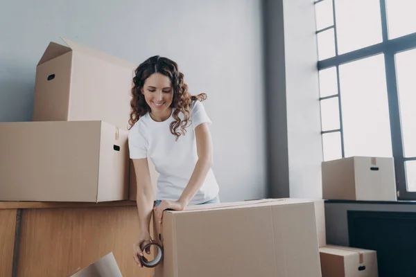 Happy girl moves alone. Young woman is packing boxes with adhesive tape. Lady is wrapping cardboard boxes with packing tape. Moving service worker preparing boxes for shipping and storage.