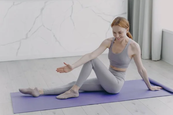 Slim sporty european girl is stretching on mat. Woman is practicing gymnastics. Concept of fitness classes at home. Sport rehabilitation. Coronavirus quarantine and healthy lifestyle.