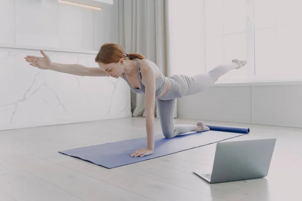 Young european woman enjoying yoga by video lessons and stretching hand. Posture exercise. Internet learning with computer. Gymnastics at home classes. Morning exercise and sport routine concept.