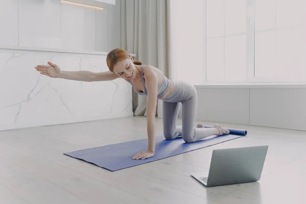 Young woman practicing yoga by video tutorial. European girl is looking at laptop screen. Posture exercise. Concept of internet learning and distance home classes on quarantine.