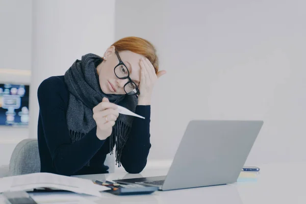 Tired overworking executive caught cold. Measuring temperature. Sad young white woman is sick. Unwell lady girl is working in office in scarf. Worker feels ill and has fever.