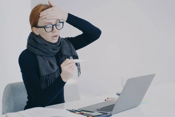 Sick manager at online conference. Young white woman got disease in office. Unwell girl is working in scarf holding thermometer and shocked with her temperature. Worker feels ill and has fever.