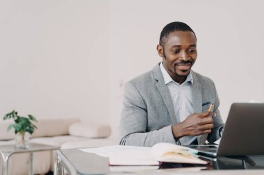 Smiling african american man working at laptop online at office desk. Black male businessperson looking at computer screen, reading email with good news about profitable deal or job promotion.