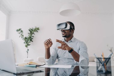 African american businessman using virtual reality glasses sitting at laptop at workplace in office. Modern black guy businessperson wearing VR goggles. Augmented Reality. Future high-tech.