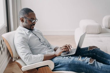 Online freelance job. Handsome african american man in front of pc. Relaxed happy businessman in formal wear is sitting in cosy chair at his apartment. Startup project, remote work.