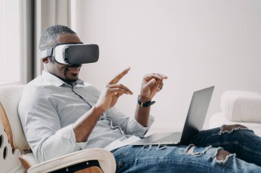African american businessman wearing VR glasses interacting with virtual reality sitting in armchair at laptop. Black guy testing modern products in cyberspace. Hightech, future technology concept.