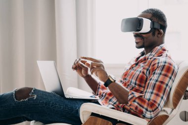 African american man in casual wear is businessman. Guy in vr headset at home office. Relaxed freelancer sitting in cosy chair and working on project on pc. High tech digital technology and vr gaming.