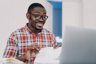 Funny african american man wearing glasses working at laptop grimaces, makes big eyes, realizing missing deadline. Black guy office worker makes faces understanding forgot business meeting.