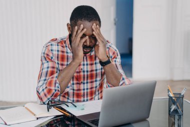 Exhausted african american man suffer headache after working at laptop. Tired frustrated black guy holds head by hands feel migraine pain at workplace. Stress at work, occupational burnout.