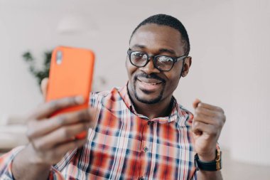 African american man wearing glasses holding smartphone having video call conversation, gesturing, takes selfie. Happy black guy looking at mobile phone smiling reading good news message.
