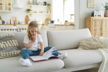 Cute caucasian little girl preschooler reading fairytale sitting on cozy couch relaxing at home alone, kid daughter reads book, interesting story in living room. Child education concept