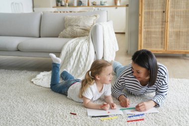 Happy family little child girl, young mother, drawing painting by colorful pencils lying together on warm floor in living room. Babysitter teaches kid to draw, develops creativity. Educational pastime
