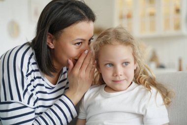 Female babysitter or mother whispering sharing secret with small kid daughter. Happy mom whisper in girl's ear, telling funny story to child, enjoying friendship, trust. Trusting family relationships.