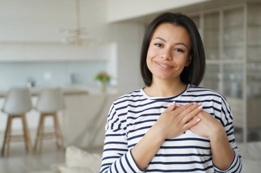 Hispanic woman keeps hands close to heart. Conceptual portrait of young happy woman showing love. Positive trendy girl in sailor shirt at home. Advertising banner mockup with copyspace.