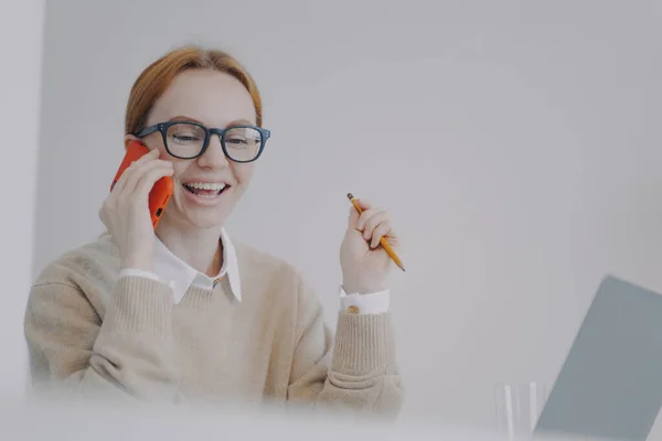 Young business woman is talking on telephone in office. Happy girl talking on cell phone to friend and laughing. Phone conference. Information and communication concept.