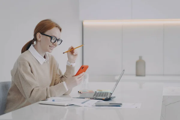 Lazy young woman is texting at workplace in office. Employee is sending messages sitting at her desk. Manager in glasses and forman wear surfing internet, having fun. Being distracted at work concept.