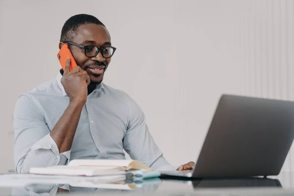 African american businessman talks on phone at laptop in office. Black male manager wearing glasses consulting client, having pleasant conversation with colleague, discussing solving problem distantly