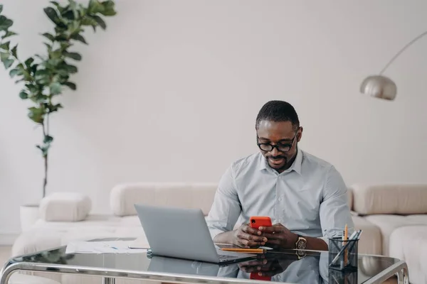 African american businessman looking at smartphone screen, sitting at laptop, working on online project, financial statistics. Black businessperson messaging with business partner by phone at office.
