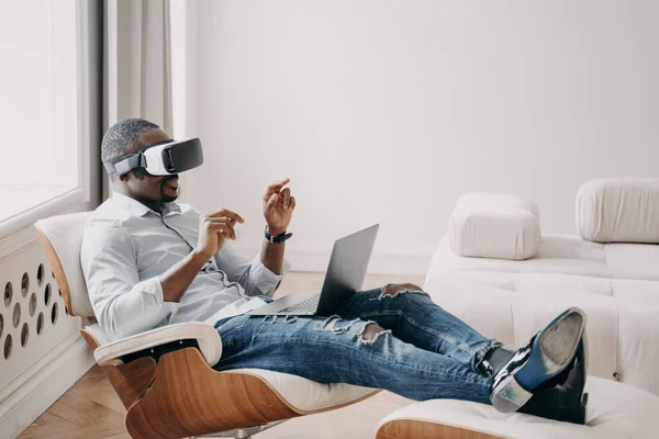 African businessman wearing VR glasses interacting with 3d objects in augmented virtual reality. Black guy using modern gadget for working in cyberspace sitting in armchair at laptop. Future high tech