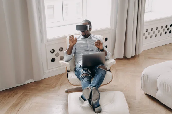 African american man in VR glasses working in augmented reality, sitting in armchair with laptop at home, pointing at virtual reality object, interacting with cyberspace, using modern high-tech device