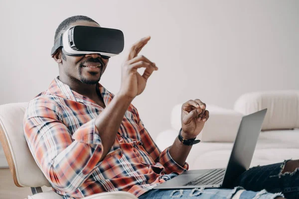Distant work and futuristic gaming. African american man in vr goggles working with laptop at home. Relaxed freelancer clicks virtual buttons. Internet technology for business and entertainment.