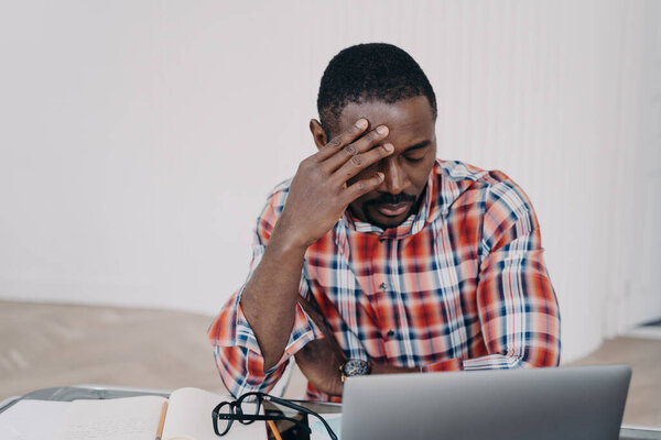 Sad tired online teacher or businessman. Afro man has online conference on laptop or working on project late. Tutor is exhausted from work. Remote study, distance learning. Deadline concept.