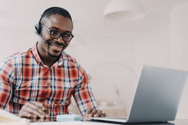 Smiling african american employee wearing headset working at laptop, looking at screen, answering a video call, communicates with customer. Black guy call center operator consultanting client.